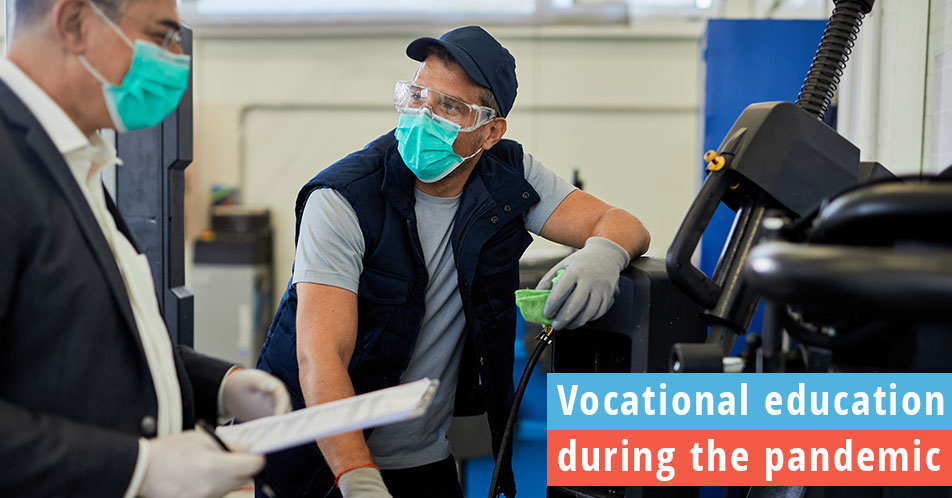 Vocational education teacher in mask while talking to student working on a car